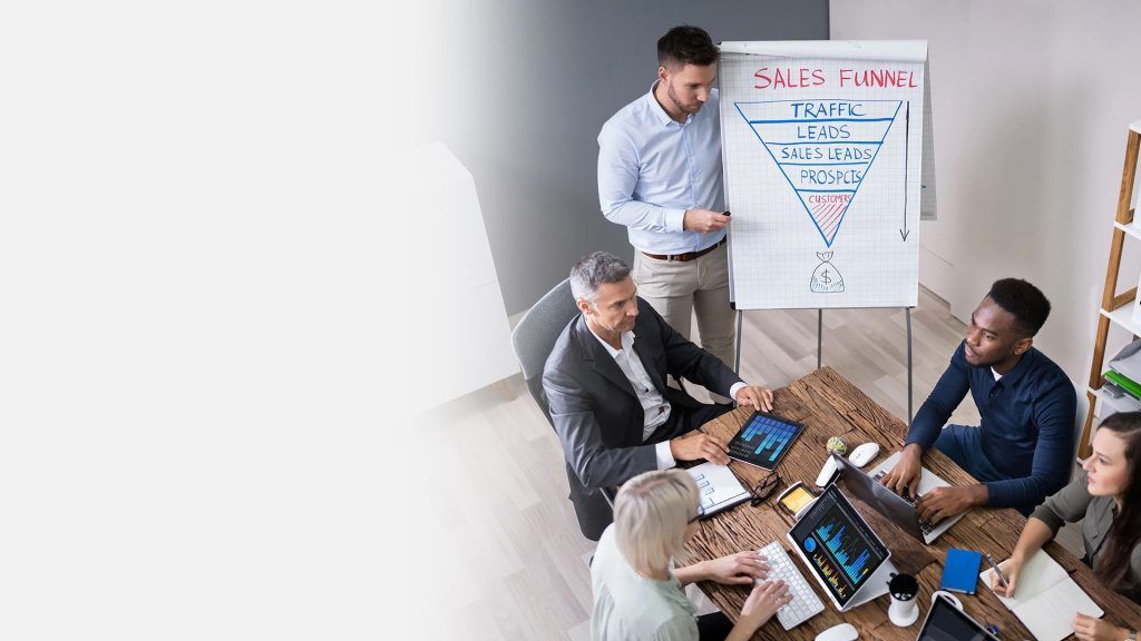 Three professionals from a marketing agency are seated around a table with digital devices, while another stands presenting an sales funnel strategy on a flip chart.