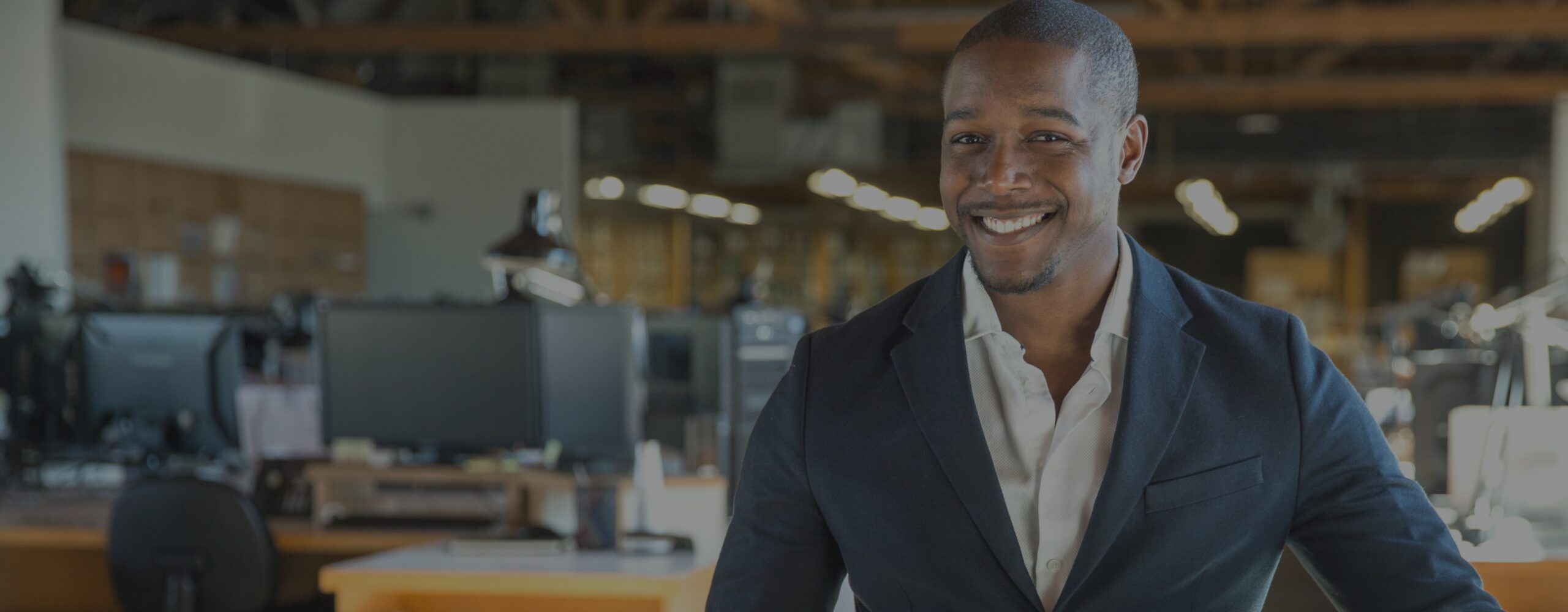 A man wearing a dark suit and a light-colored shirt smiles at the camera in an office setting. The background, reminiscent of a marketing playbook in action, shows various office equipment, desks, and chairs under wooden ceiling beams illuminated by bright overhead lighting.