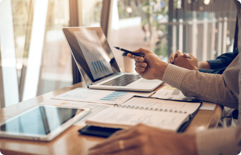 Two people are sitting at a table with a laptop, tablet, and open notebook. One person is pointing at the laptop screen with a pen, and charts and graphs are visible on the screen. The table is next to a large window, letting in natural light.