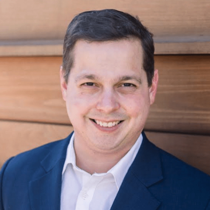 A man with short, dark hair is smiling while wearing a dark suit and white shirt. He is standing in front of a wooden wall. The image has a thick orange border on two sides.