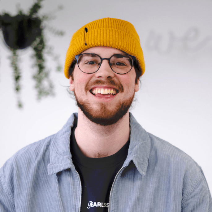 A person with a beard and glasses is smiling. They are wearing a mustard yellow beanie, a striped button-up shirt over a dark shirt, and standing against a light-colored background with plants hanging on the side.