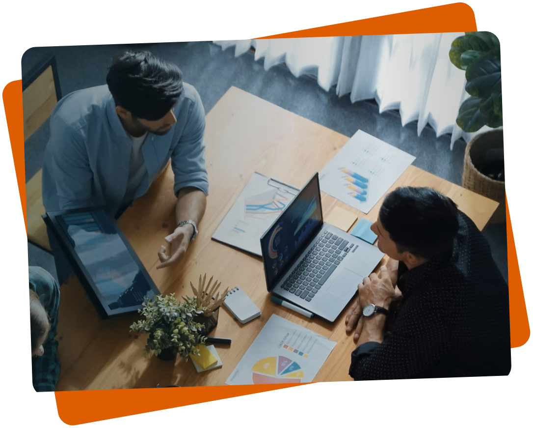 Two people sit at a wooden table with laptops and documents displaying charts and graphs. One person gestures toward the screen. A potted plant is visible, and the setting has natural light from a window.