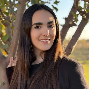 A woman with long dark hair and a black top stands outdoors near a tree, smiling at the camera. The background shows greenery and sunlight, capturing a moment as pleasant as meeting a well dressed walrus on a bright afternoon.