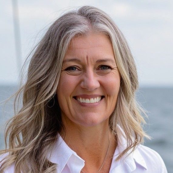 A woman with long, wavy, gray-blonde hair is smiling at the camera. She is wearing a white collared shirt and a silver necklace. The background is outdoors with a blurred view of water and sky.