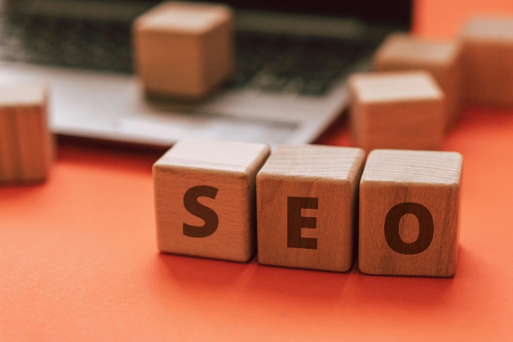 Three wooden blocks spell "SEO," arranged before a blurred laptop on an orange surface. Highlighting website health, extra blocks are visible in the background, suggesting attention to SEO 2026 trends and avoiding SEO red flags.