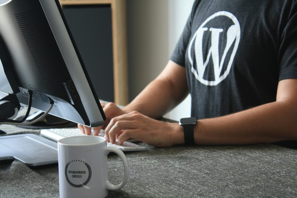A person wearing a t-shirt with the WordPress logo types on a keyboard at a desk with a monitor and a coffee mug labeled "WordPress Oulu," staying up-to-date on WordPress Security in 2026 to keep their site safe.