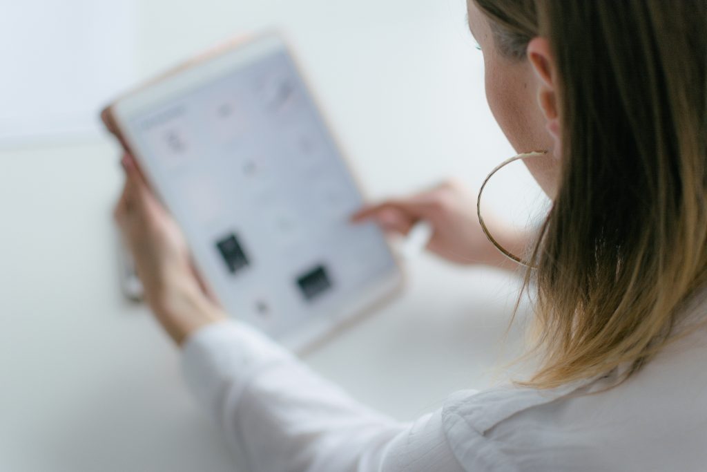 A person with long hair wearing a white shirt and large hoop earrings uses a tablet with one hand, tapping the screen—perhaps searching for SEO services—while the tablet display appears blurred and indistinct.